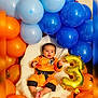baby, orange_outfit, blue_balloons, orange_balloons, gold_balloon, number_3, dragon_ball_z, figurines, fluffy_blanket, birthday, celebration, child, smiling, lying_down, indoor, cute, colorful, toy_figures, decorations, happy