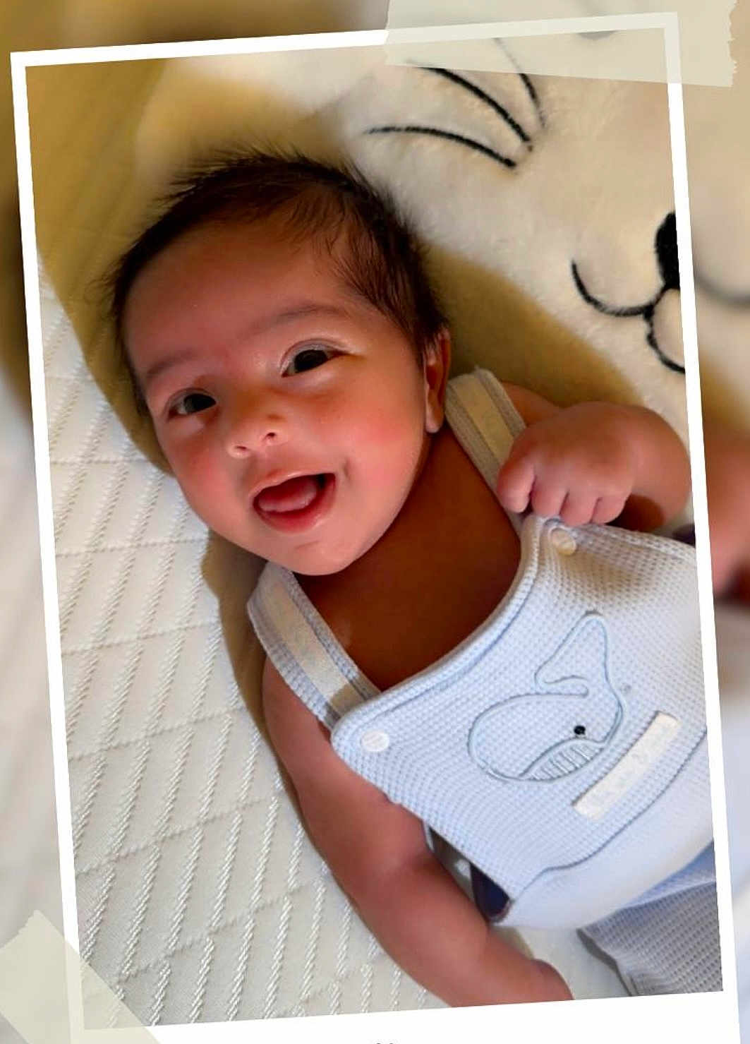 Hans is registered to the contest to win money with this photo: baby, smiling, infant, cute, happy, lying_down, white_background, blue_clothing, whale_embroidery, raised_fist, closeup, portrait, child, soft_texture, textured_surface, tape_frame, bright_eyes, adorable, indoor, sleepwear