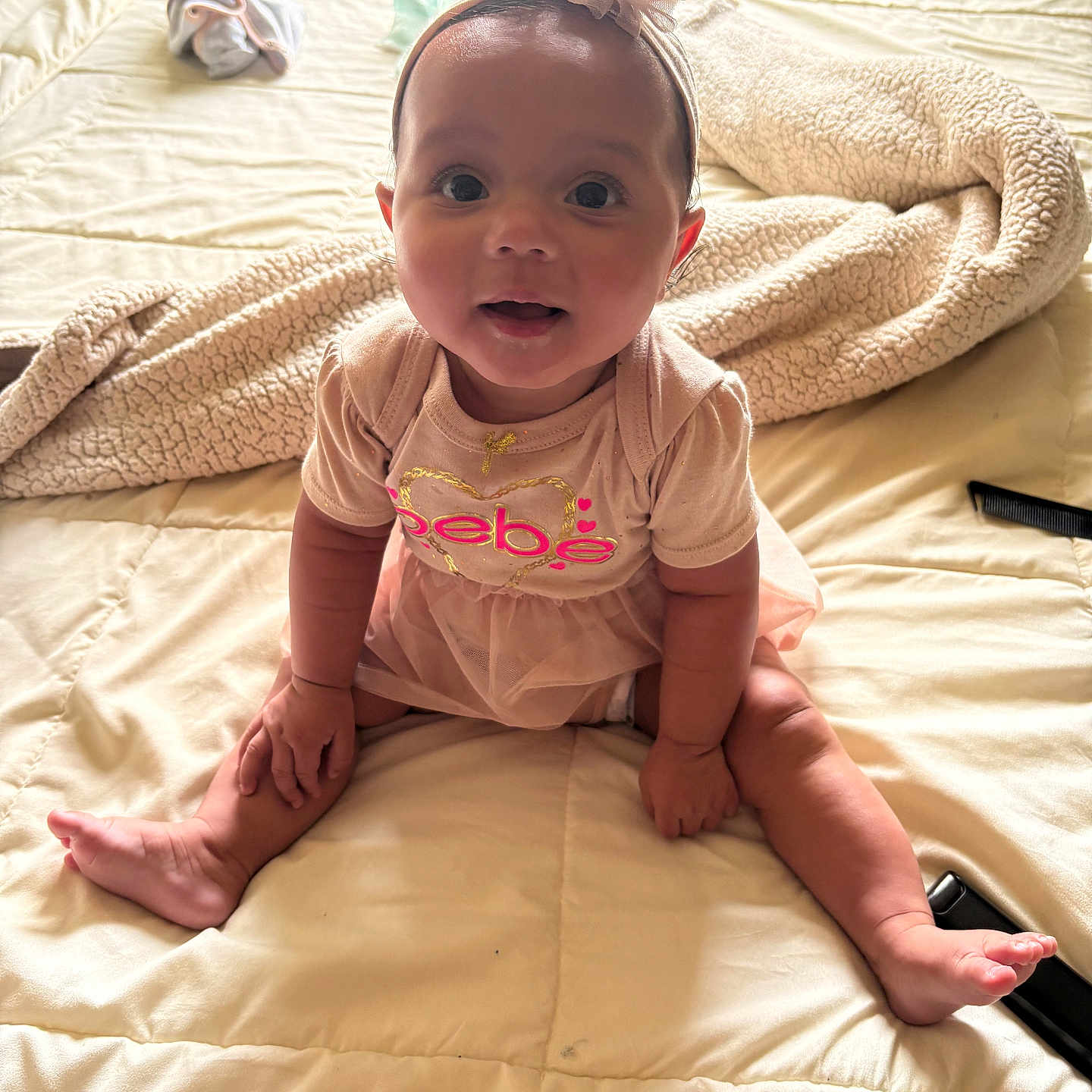Kali is registered to the contest to win money with this photo: baby, bed, blanket, bow, child, curious, cute, face, footwear_absent, hand, headband, indoors, infant, person, pink_dress, sitting, skin, smiling, soft_lighting, toes