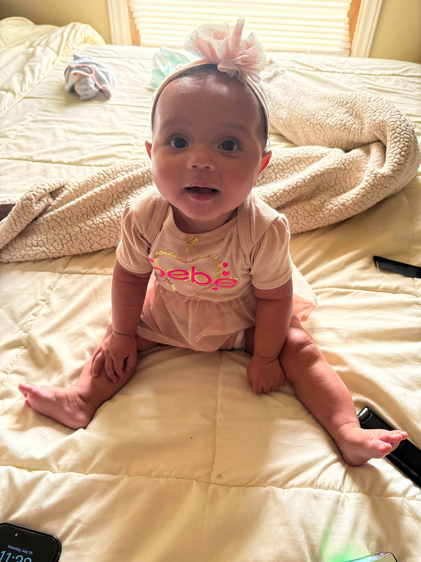 Kali is registered to the contest to win money with this photo: baby, child, infant, bed, blanket, pink_dress, headband, bow, smiling, sitting, indoors, cute, curious, soft_lighting, person, footwear_absent, skin, hand, face, toes