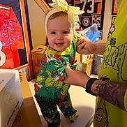 Paisley is registered to the contest to win money with this photo: baby, child, pajamas, headband, yellow_bow, table, tattoo, adult_hand, sports_memorabilia, framed_jersey, framed_photos, indoor, person, smiling, colorful_clothing, green_pajamas, foot, holding_hands, wall_art, bright_colors