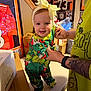 baby, child, pajamas, headband, yellow_bow, table, tattoo, adult_hand, sports_memorabilia, framed_jersey, framed_photos, indoor, person, smiling, colorful_clothing, green_pajamas, foot, holding_hands, wall_art, bright_colors