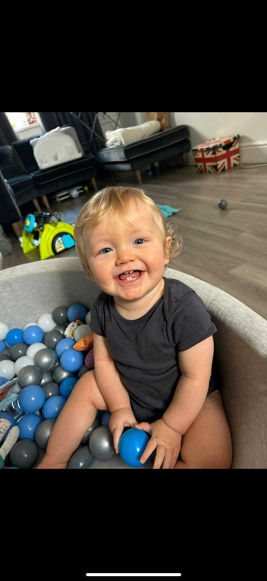 Milo-Chrisopher is registered to the contest to win money with this photo: toddler, child, ball_pit, blue_ball, smiling, blonde_hair, blue_eyes, black_onesie, indoor, living_room, sofa, toy, floor, happy, person, play, furniture, home, cute, baby