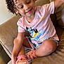 casual_clothing, child, couch, cross_legged, curly_hair, cute, furniture, happy, home, indoor, person, pink_shirt, playful, relaxed, seated, shorts, smiling, sneakers, socks, toddler