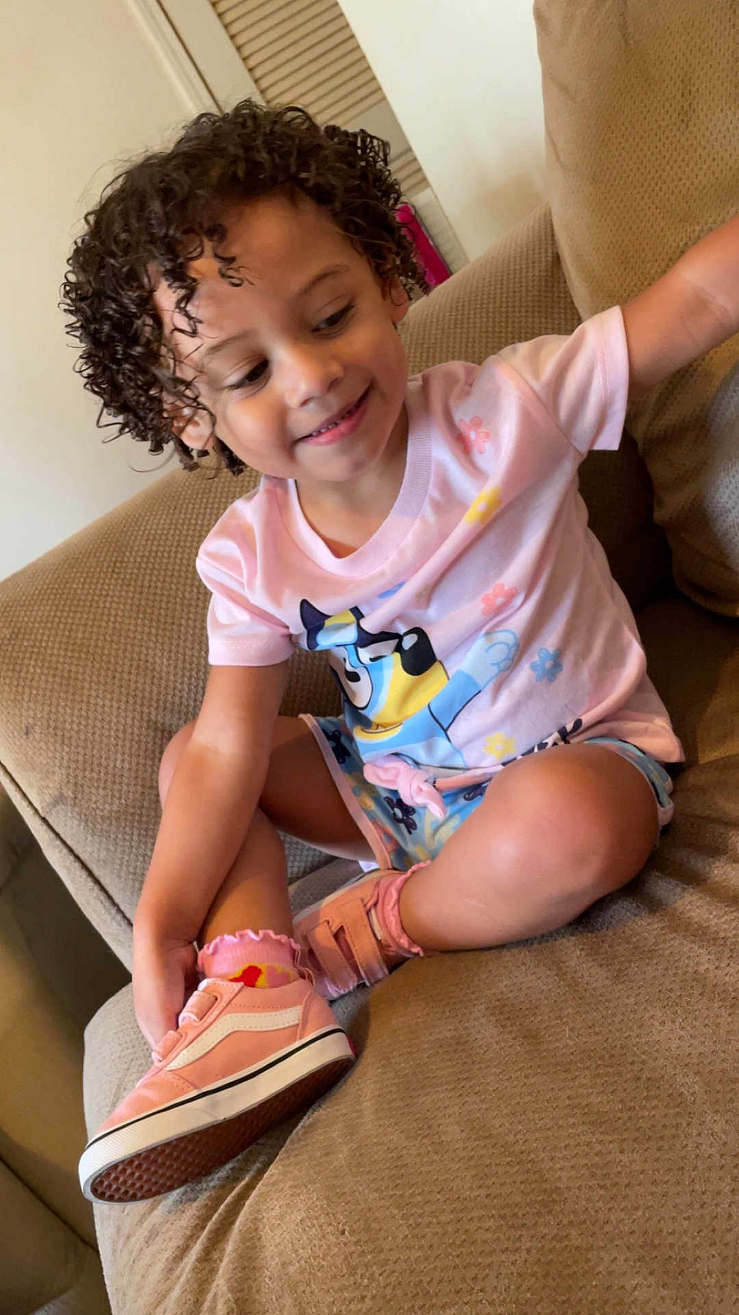 Skylet joined the competition — help win amazing prizes! toddler, child, curly_hair, pink_shirt, shorts, sneakers, socks, couch, indoor, smiling, happy, casual_clothing, seated, cross_legged, cute, playful, person, furniture, home, relaxed