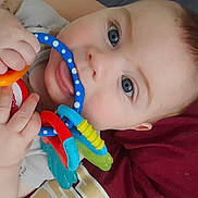 Paxton is registered to the contest to win money with this photo: baby, blue_eyes, chewing, child, close_up, colorful, cute, face, fingers, hand, indoors, infant, maroon_blanket, play, portrait, resting, skin, soft_clothing, teething_toys, young