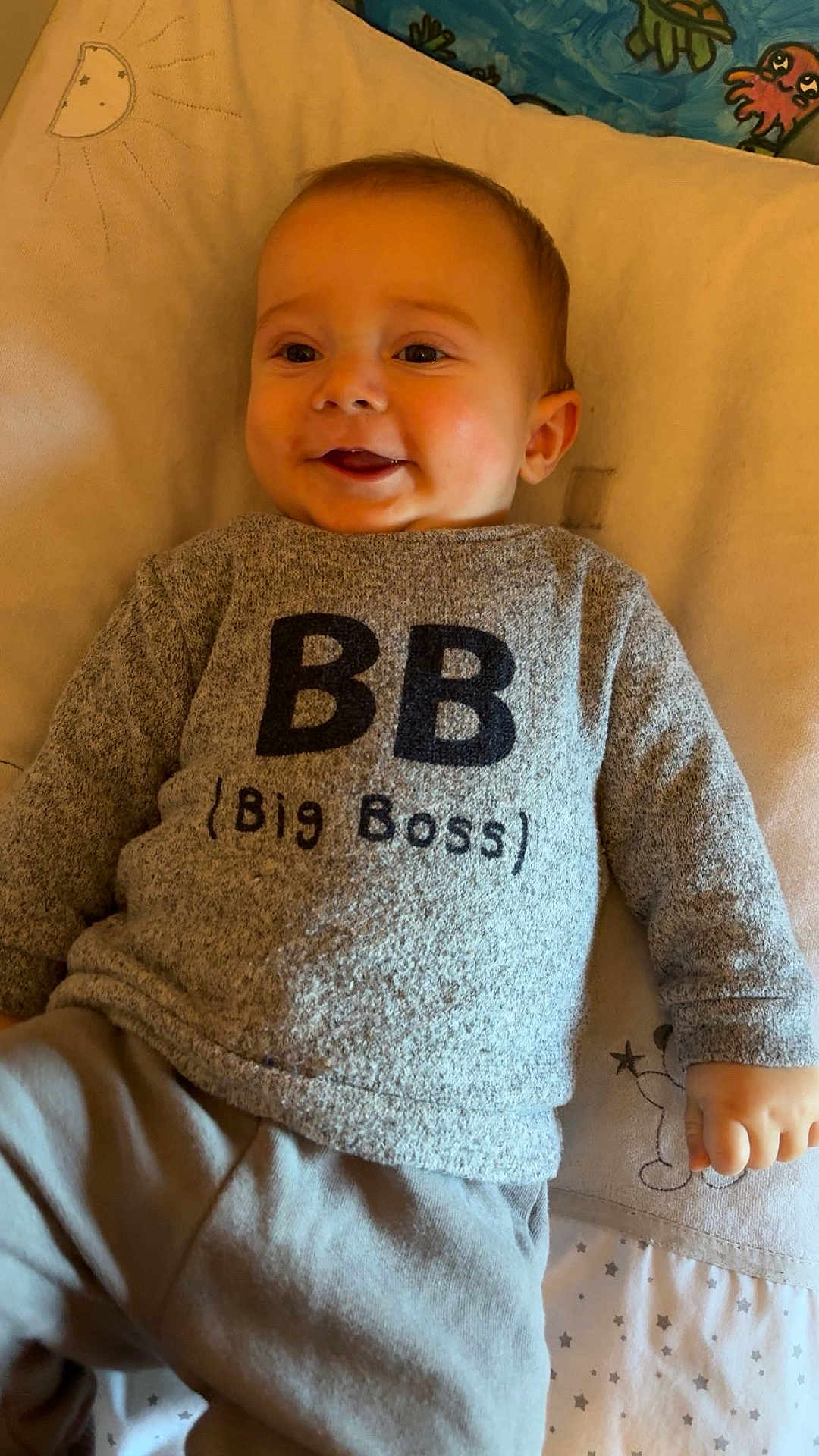 Soan participe au concours pour gagner de l'argent avec cette photo : baby, smiling, sweater, grey_clothing, lying_down, bed, pillow, cute, child, infant, face, hand, text_on_clothing, indoor, soft_lighting, blanket, cozy, happy, person, portrait