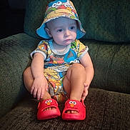 Michael is registered to the contest to win money with this photo: toddler, child, hat, bucket_hat, red_slippers, elmo_slippers, colorful_clothing, cartoon_print, sitting, couch, green_couch, indoor, serious_expression, baby, footwear, person, cute, portrait, young_child, home