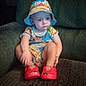 toddler, child, hat, bucket_hat, red_slippers, elmo_slippers, colorful_clothing, cartoon_print, sitting, couch, green_couch, indoor, serious_expression, baby, footwear, person, cute, portrait, young_child, home