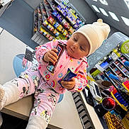 Muna is registered to the contest to win money with this photo: toddler, child, store, counter, candy_bar, snacks, pajamas, beanie, indoor, shopping, convenience_store, shelf, colorful_clothing, cute, person, holding, curious, floor, footwear, toy