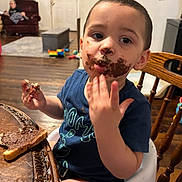 Landon is registered to the contest to win money with this photo: toddler, child, chocolate, face_paint, high_chair, table, bread, food, messy, indoor, wooden_floor, person, furniture, toy, living_room, blue_shirt, hand, smile, cute, young