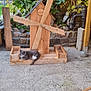 cat, wooden_windmill, garden, plants, stone_wall, outdoor, concrete_floor, rustic, greenery, wood, relaxing, pet, animal, feline, nature, shrubbery, decor, resting, peaceful, daylight