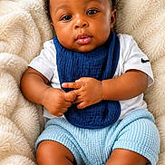 Ishmael is registered to the contest to win money with this photo: baby, infant, child, curly_hair, blue_bib, white_shirt, blue_shorts, grey_socks, fluffy_blanket, soft_texture, cute, portrait, indoors, comfortable, relaxed, chubby_arms, chubby_legs, looking_at_camera, skin, hands
