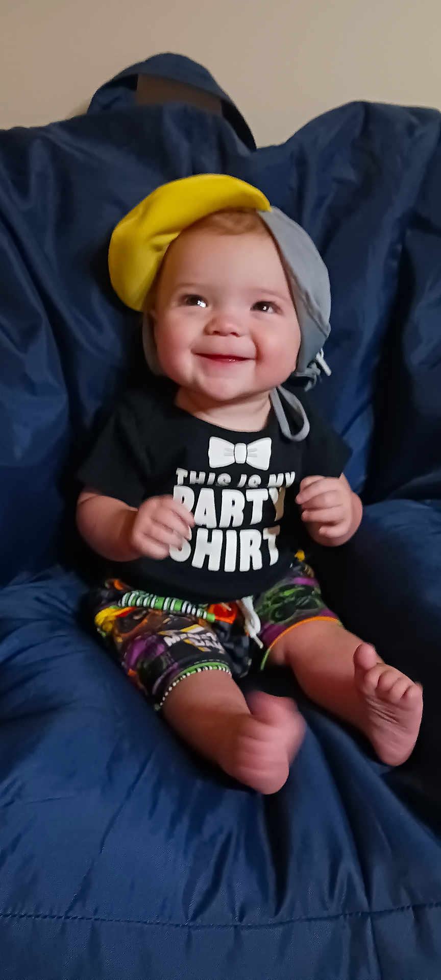 Ethan is registered to the contest to win money with this photo: baby, smile, hat, black_shirt, party_shirt, colorful_shorts, blue_chair, cushion, happy, child, indoors, cute, sitting, feet, hands, portrait, infant, clothing, headwear, person