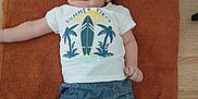 Flavio participe au concours pour gagner de l'argent avec cette photo : baby, child, toddler, curly_hair, tshirt, denim_shorts, brown_towel, wooden_floor, smiling, lying_down, infant, baby_care, face, person, indoor, clothing, happy, skin, hand, foot