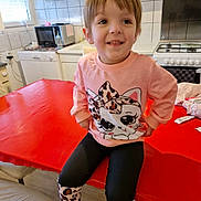 Angelina a rejoint le concours — aidez-le/la à gagner de superbes lots ! child, smiling, pink_sweater, cat_design, leopard_print, leggings, sitting, red_table, kitchen, stove, microwave, window, dishwasher, white_cabinet, floor, chair, person, happy, casual_clothing, indoor