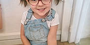 Angelina a rejoint le concours — aidez-le/la à gagner de superbes lots ! child, girl, glasses, pigtails, smiling, indoor, floor, denim_overalls, white_tshirt, happy, portrait, cute, young, casual_clothing, person, wooden_floor, home, small_child, sitting, playful