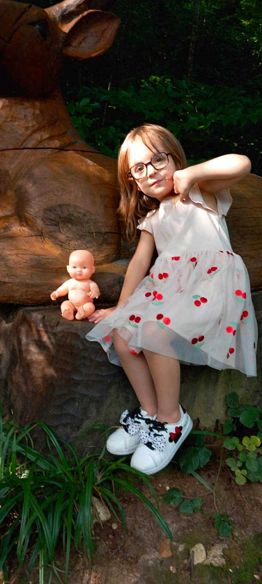 Angelina participe au concours pour gagner de l'argent avec cette photo : child, girl, glasses, dress, cherries, white_shoes, doll, wooden_sculpture, outdoor, nature, greenery, plants, tree, playful, cute, portrait, sitting, casual, sunlight, young