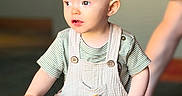 Atticus is registered to the contest to win money with this photo: baby, child, toddler, portrait, indoor, standing, striped_shirt, overalls, curious, face, eyes, mouth, table, snacks, food, adult, background, warm_lighting, closeup, photography