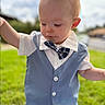 baby, child, outdoor, portrait, face, bow_tie, vest, dress_shirt, plaid, formalwear, blond_hair, blue_clothing, standing, arms_raised, grass, garden, sunlight, blurred_background, smiling, fashion