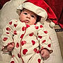 baby, infant, santa_hat, onesie, red_hearts, cushion, soft_toy, holiday, festive, cute, portrait, indoor, child, face, clothing, cozy, smile, holiday_season, young, celebration