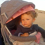 Lya participe au concours pour gagner de l'argent avec cette photo : toddler, child, stroller, blanket, curly_hair, blue_eyes, outdoor, beach, sunset, clothing, person, portrait, infant, baby, warm_light, seated, comfort, expression, soft_texture, casual