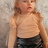child, girl, portrait, curly_hair, blue_eyes, beige_top, black_skirt, buttons, indoor, soft_lighting, fashion, cute, young, person, toddler, background_pattern, long_sleeves, expression, standing, photography