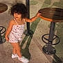 casual_clothing, child, concrete_floor, curly_hair, cute, footwear, holding, legs, nighttime, outdoor, person, playground, recreation, shadow, small_person, smiling, standing, white_shoes, wooden_stool, young_child
