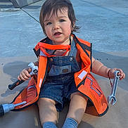 Jacob is registered to the contest to win money with this photo: toddler, child, playground, orange_vest, denim_overalls, toy_tools, wrench, hammer, socks, outdoor, concrete, bench, play_equipment, sunlight, hair, face, person, sitting, cute, playtime