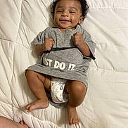Jasper is registered to the contest to win money with this photo: baby, infant, smiling, curly_hair, diaper, grey_tshirt, just_do_it, blanket, bed, fist, happy, lying_down, skin, cute, playful, person, wristband, arm, indoors, child