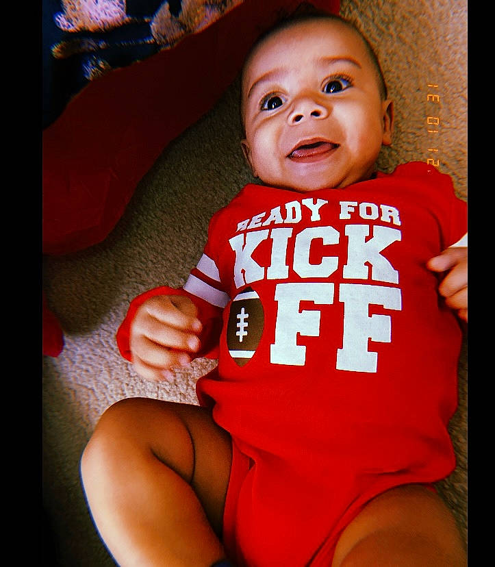 Garrett is registered to the contest to win money with this photo: baby, baby_toddler_clothing, comfort, elbow, finger, gesture, happy, human_leg, jersey, lip, muscle, neck, orange, person, sleeve, sportswear, t_shirt, thigh, thumb, toddler