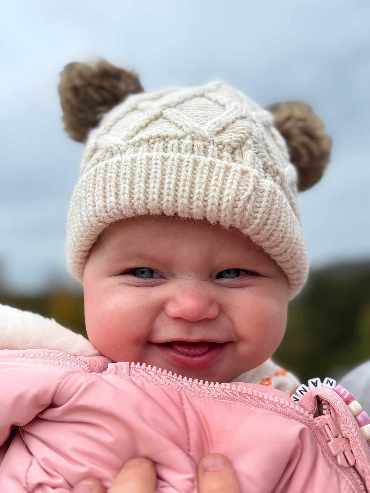 Mabel is registered to the contest to win money with this photo: baby, child, smiling, hat, knit_hat, pom_poms, pink_jacket, outdoor, cold_weather, cute, face, cheeks, happy, person, portrait, winter_clothing, closeup, hand, zipped_jacket, blue_eyes