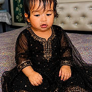 Avleen is registered to the contest to win money with this photo: baby, child, infant, sitting, portrait, indoors, bedroom, bed, bedspread, black_dress, gold_embroidery, formal_clothing, curly_hair, dark_hair, looking_down, face, hands, fashion, furniture, colorful_background