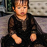 baby, bed, bedroom, bedspread, black_dress, child, colorful_background, curly_hair, dark_hair, face, fashion, formal_clothing, furniture, gold_embroidery, hands, indoors, infant, looking_down, portrait, sitting