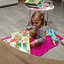 Maëlys a rejoint le concours — aidez-le/la à gagner de superbes lots ! book, books, bottle, casual_clothing, child, cozy, doll, floor, girl, hair_clip, home, indoor, pink_blanket, play, reading, sitting, stuffed_animal, table, tiled_floor, toy