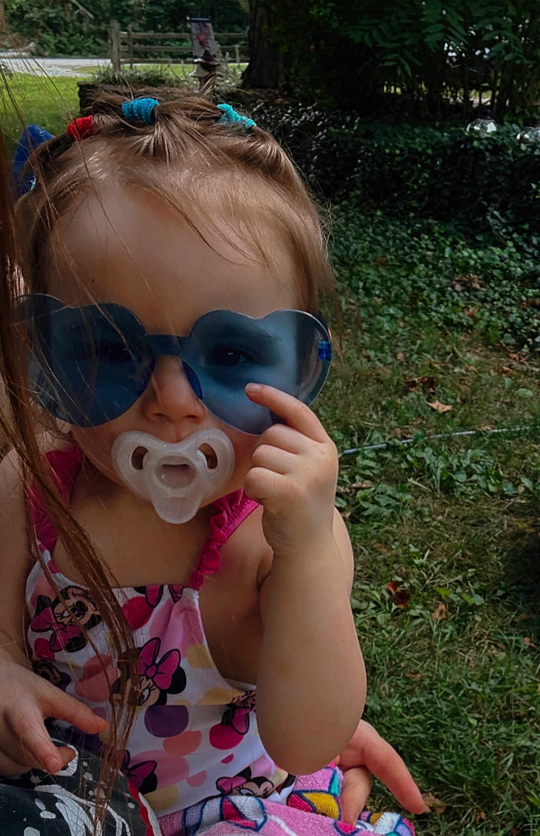 Tessa is registered to the contest to win money with this photo: toddler, child, sunglasses, pacifier, outdoor, grass, summer, hair_ties, swimsuit, minnie_mouse, hand, pointing, cute, baby, person, playful, colorful, fun, portrait, sunny