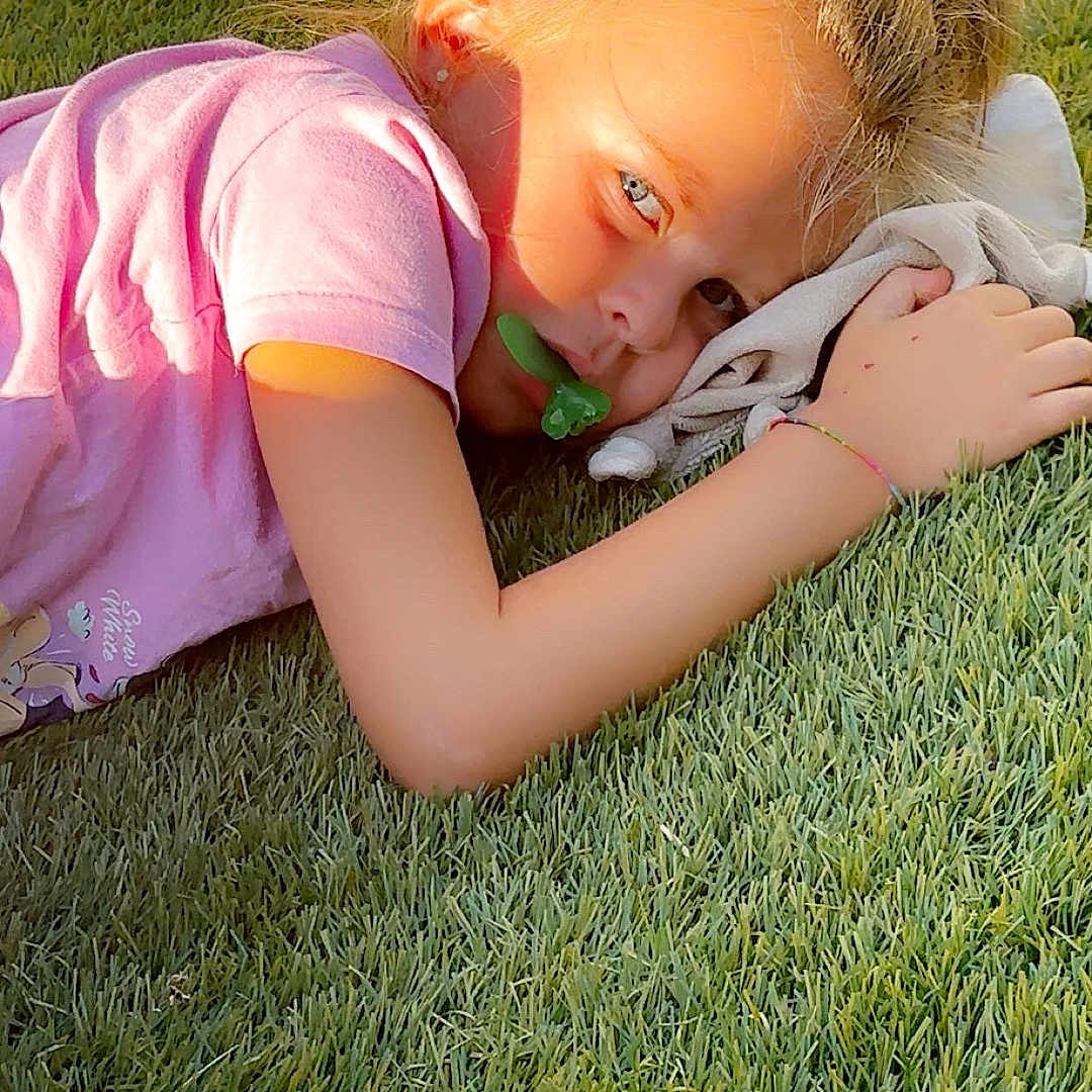 Jodie participe au concours pour gagner de l'argent avec cette photo : blonde_hair, casual, child, daylight, earrings, fabric, face, grass, hair_tie, hand, laying_down, nature, outdoor, pacifier, pink_shirt, portrait, relaxing, shadow, sunlight, youth