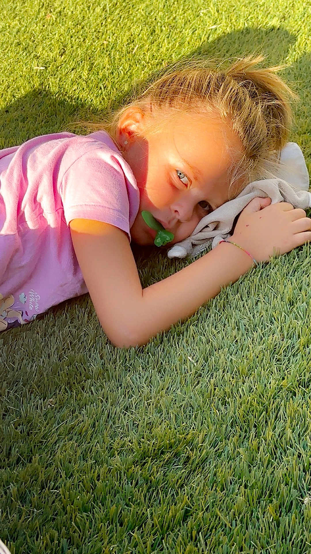 Jodie participe au concours pour gagner de l'argent avec cette photo : child, grass, pacifier, pink_shirt, sunlight, outdoor, laying_down, blonde_hair, hair_tie, hand, face, earrings, shadow, fabric, relaxing, nature, daylight, portrait, casual, youth