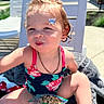 Scarlett is registered to the contest to win money with this photo: baby, blue_butterfly, casual, cereal, chair, child, curly_hair, earrings, face, happy, necklace, outdoor, person, playful, smile, snack, summer, sunlight, swimsuit, toddler