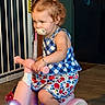 baby_gate, blue_top, bracelet, child, curly_hair, cute, floor, footwear, indoor, light, necklace, pacifier, patterned_shorts, person, pink, playtime, sitting, toddler, toy_scooter, young_child