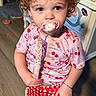 baby, blue_eyes, bracelet, cabinet, checkered_shorts, cherry_pattern, child, clothing, curly_hair, cute, floor, indoor, pacifier, person, pink_shirt, portrait, red_shorts, stickers, toddler, wood_floor