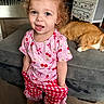 blue_eyes, bracelet, cat, checkered_pants, cherry_pattern, child, couch, curly_hair, cute, earrings, floor, furniture, ginger_cat, indoor, living_room, person, pink_shirt, smiling, standing, toddler