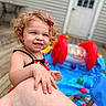 background, blur, child, curly_hair, daylight, deck, hand, happy, inflatable_pool, leg, outdoor, person, playing, red_bow, skin, smile, summer, swimsuit, toddler, toys