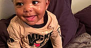 Kanan is registered to the contest to win money with this photo: toddler, child, smiling, curly_hair, mickey_mouse, shirt, black_pants, sneakers, bed, pillow, blanket, indoors, happy, person, cute, young_child, seated, home, casual_clothing, portrait