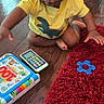 Kanan joined the competition — help win amazing prizes! toddler, child, yellow_shirt, braided_hair, toy_phone, toy_book, red_rug, wooden_floor, indoor, playtime, sitting, curious, young_child, floor_play, baby_toys, home, casual_clothing, car_seat, colorful, calm