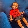 baby, child, smiling, blue_chair, red_shirt, skirt, sitting, happy, cute, indoors, plush_furniture, person, portrait, small_child, cozy, casual_clothing, barefoot, infant, expression, comfort