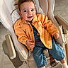 baby, child, smile, orange_jacket, denim_overalls, baby_rocker, indoor, happy, cute, infant, clothing, footwear, socks, face, person, seated, furniture, wooden_floor, playful, comfort