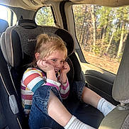 Haley is registered to the contest to win money with this photo: child, car_seat, vehicle_interior, smiling, blonde_hair, striped_shirt, jeans, white_socks, window, trees, sunlight, seatbelt, happy, casual_clothing, portrait, young, sitting, car, daylight, interior