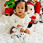 Eva Mota is registered to the contest to win money with this photo: toddler, child, white_clothing, white_socks, teddy_bear, christmas_decor, plush_toys, elf_toy, santa_toy, festive, holiday, snow_effect, indoor, cute, smiling, sitting, holiday_season, decorations, toy, happy