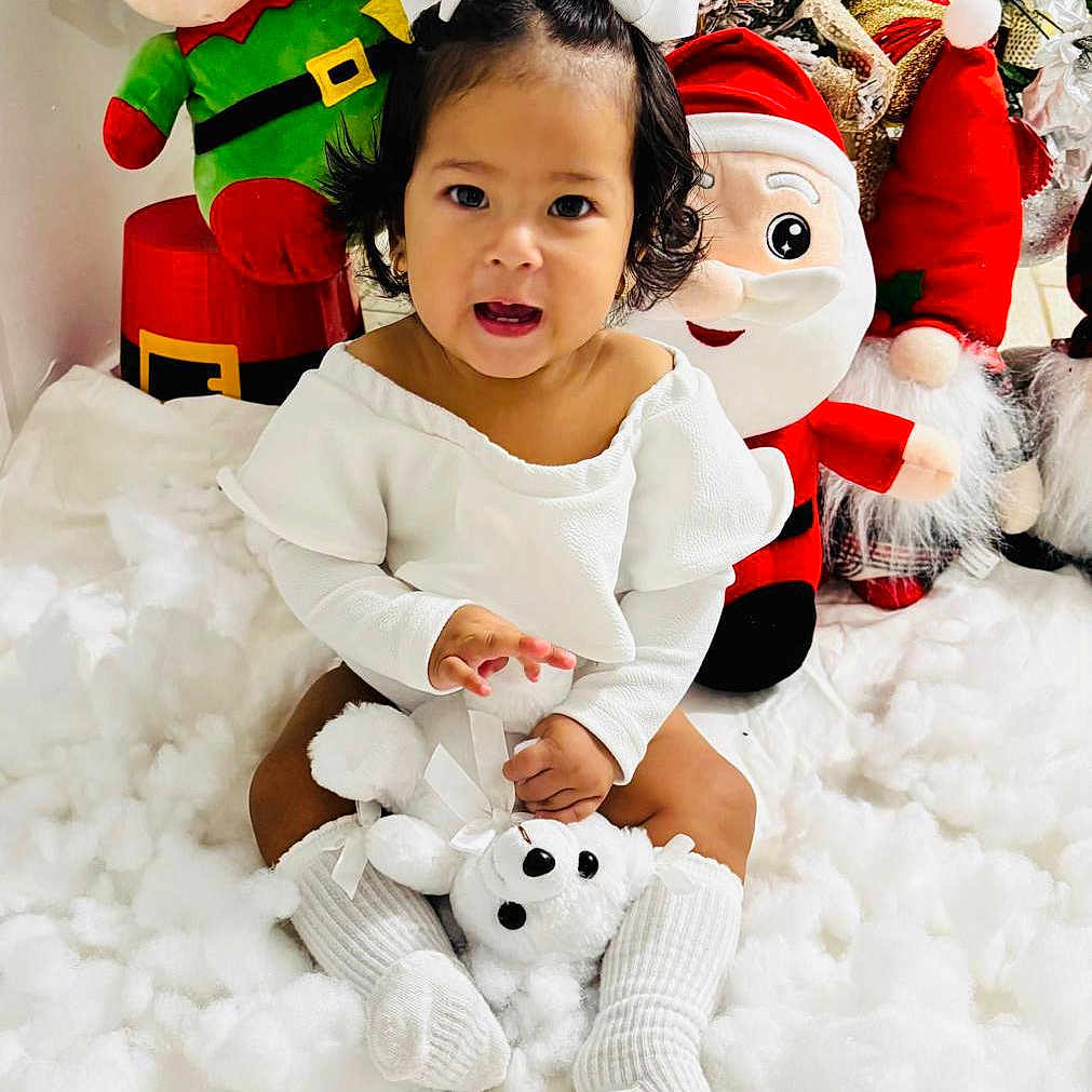 Eva Mota is registered to the contest to win money with this photo: child, christmas_decor, cute, decorations, elf_toy, festive, happy, holiday, holiday_season, indoor, plush_toys, santa_toy, sitting, smiling, snow_effect, teddy_bear, toddler, toy, white_clothing, white_socks